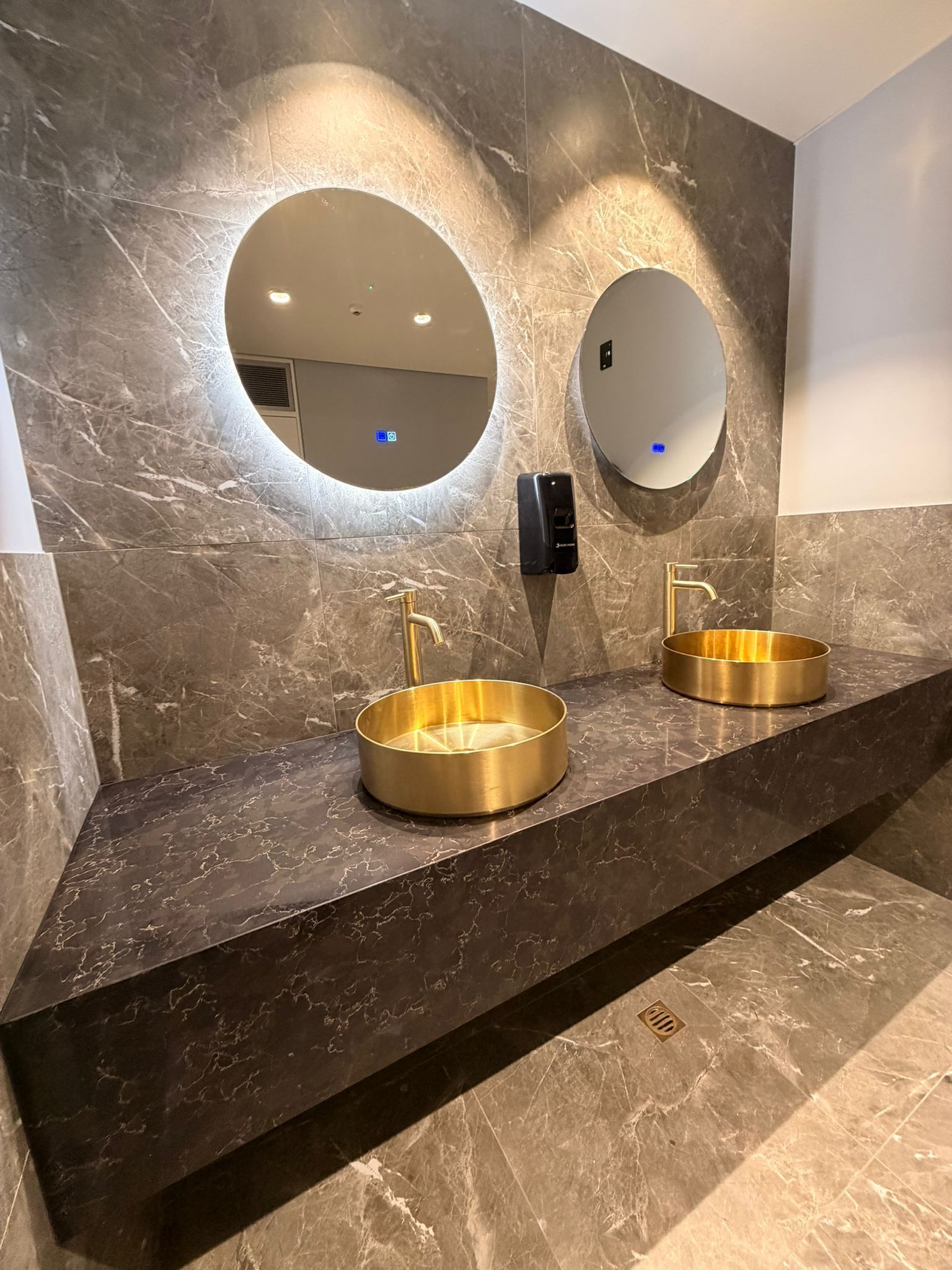 Two golden sinks on a dark countertop with round mirrors and a soap dispenser in a restroom.