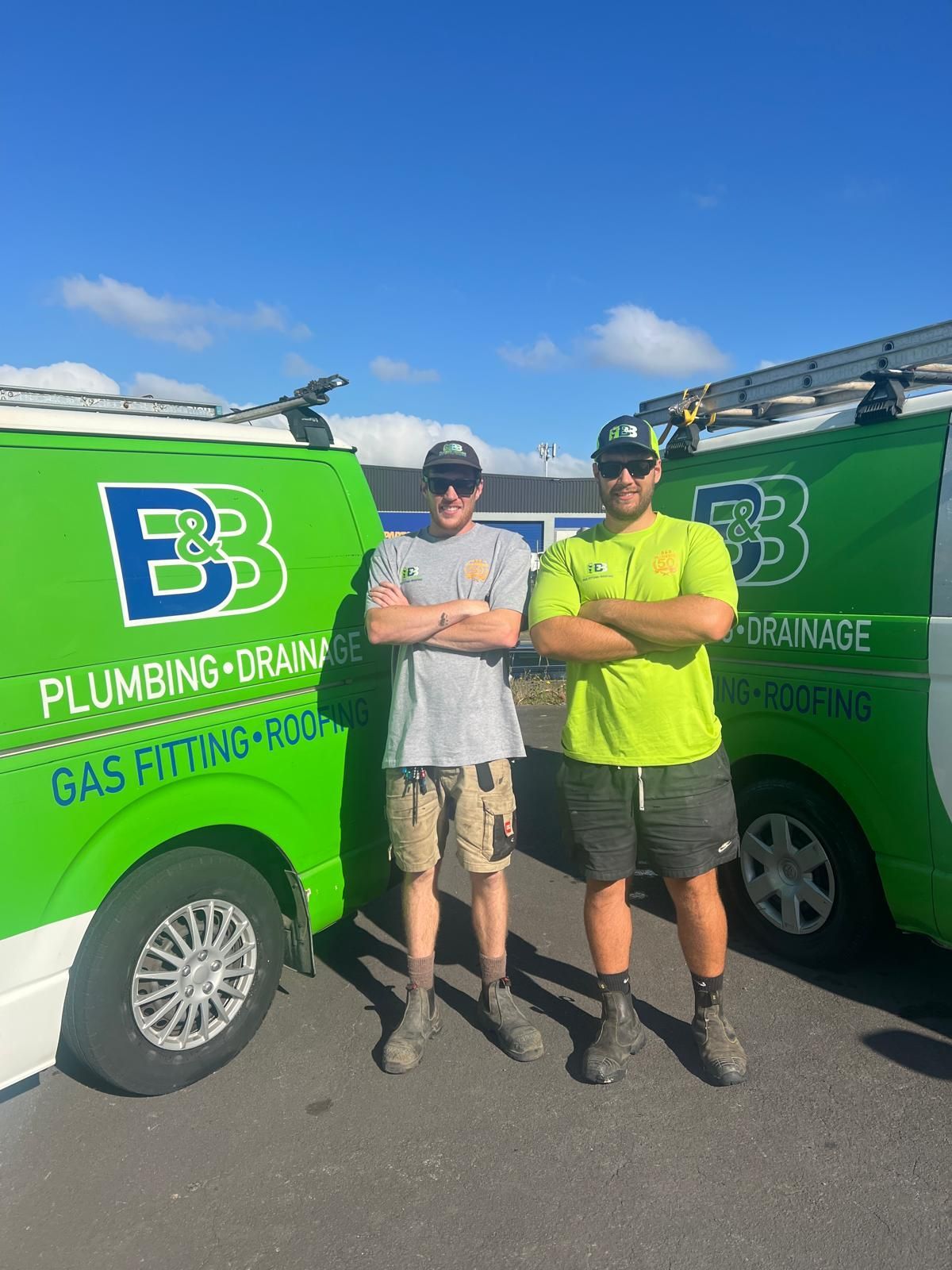 Two men standing in front of green plumbing vans. They are in work clothes, arms crossed.