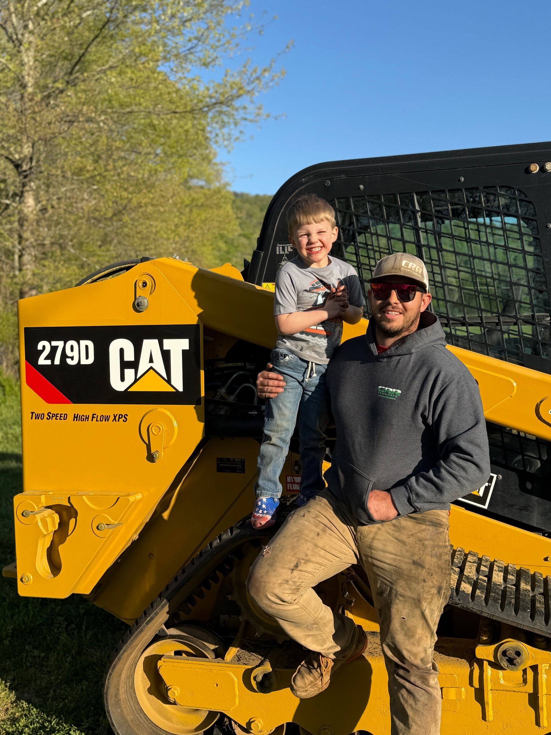 A man and a child are standing next to a yellow cat bulldozer.