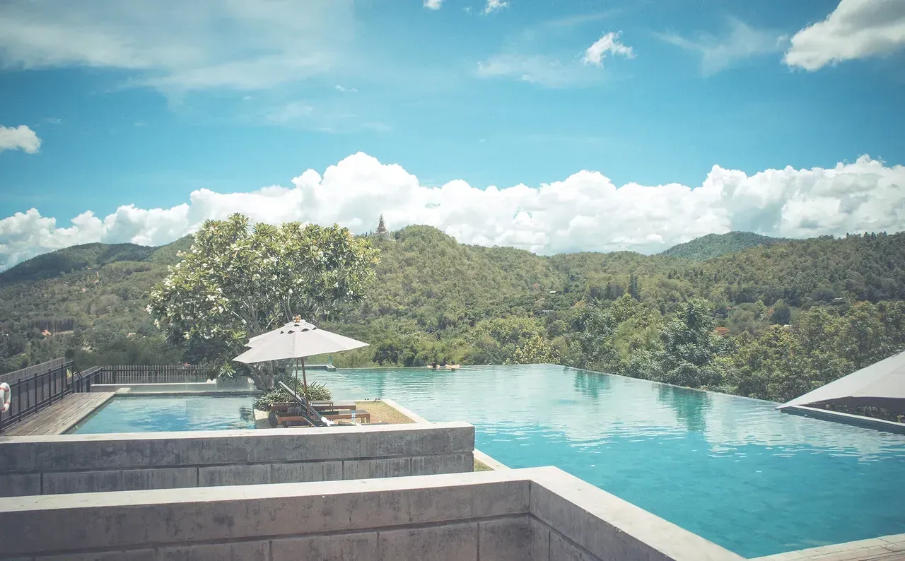 A scenic infinity pool overlooking lush green mountains under a bright blue sky with clouds.