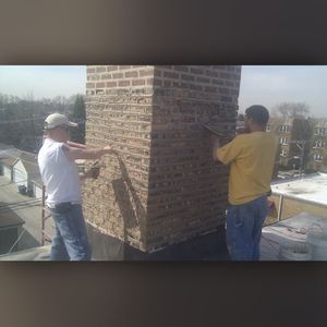 Two men are working on a brick chimney on a roof.