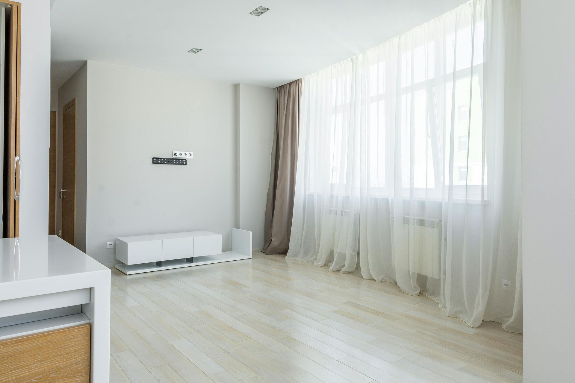 Bright, empty living room with white walls, pale wood floor, and large sheer-curtained windows