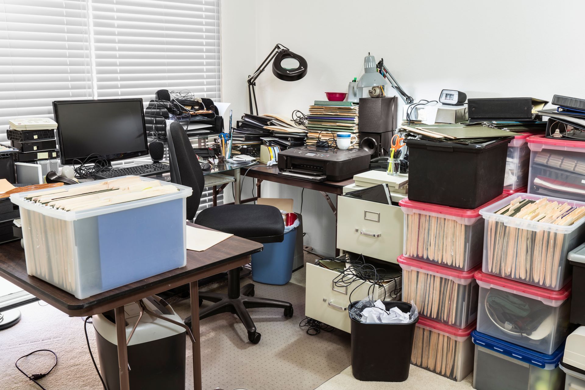 Cluttered office workspace with computer, desk lamp, filing boxes, and stacked papers