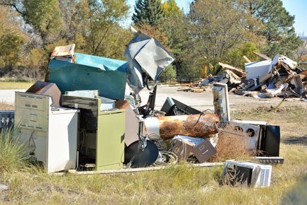 Piled old appliances and debris in a grassy roadside lot under trees
