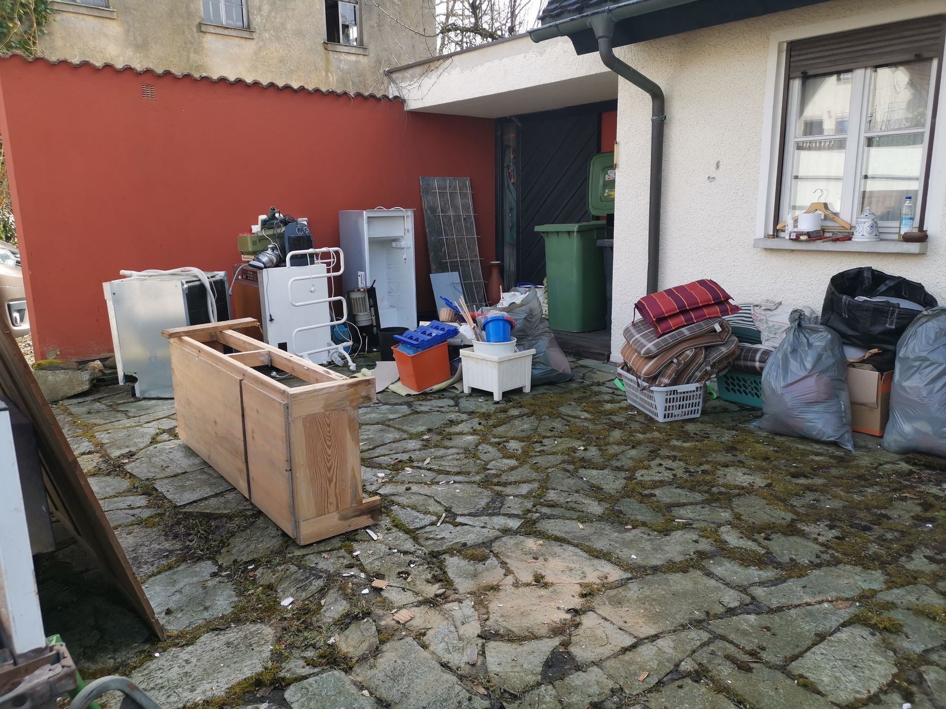 Outdoor courtyard with stacked appliances, a wooden crate, bags, and clutter against a red wall