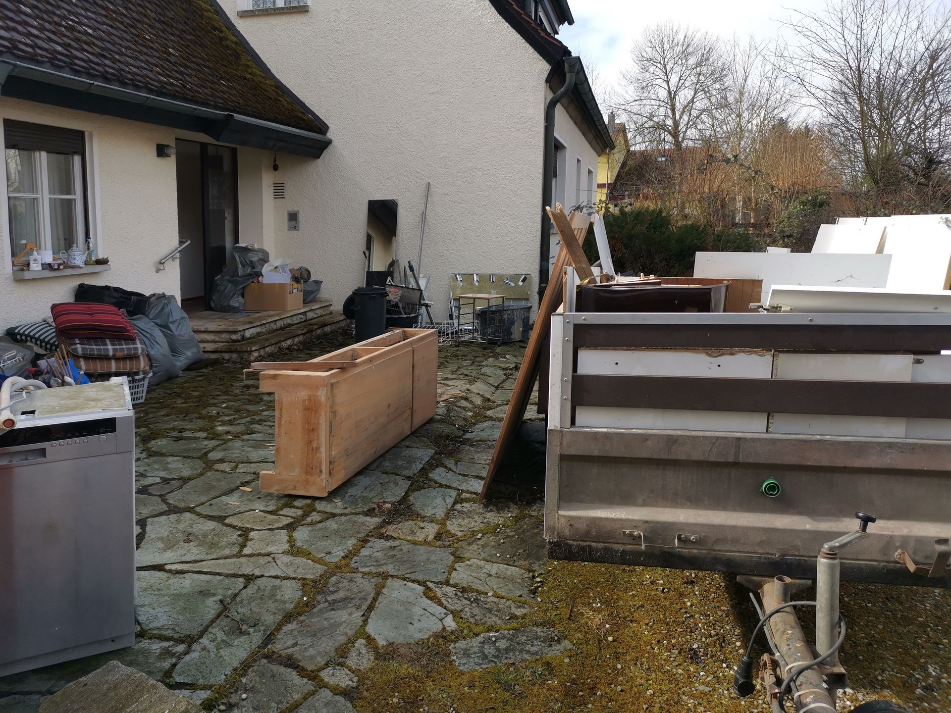 Backyard courtyard with stacked wooden crates, paving stones, and a large trailer or bin near a house
