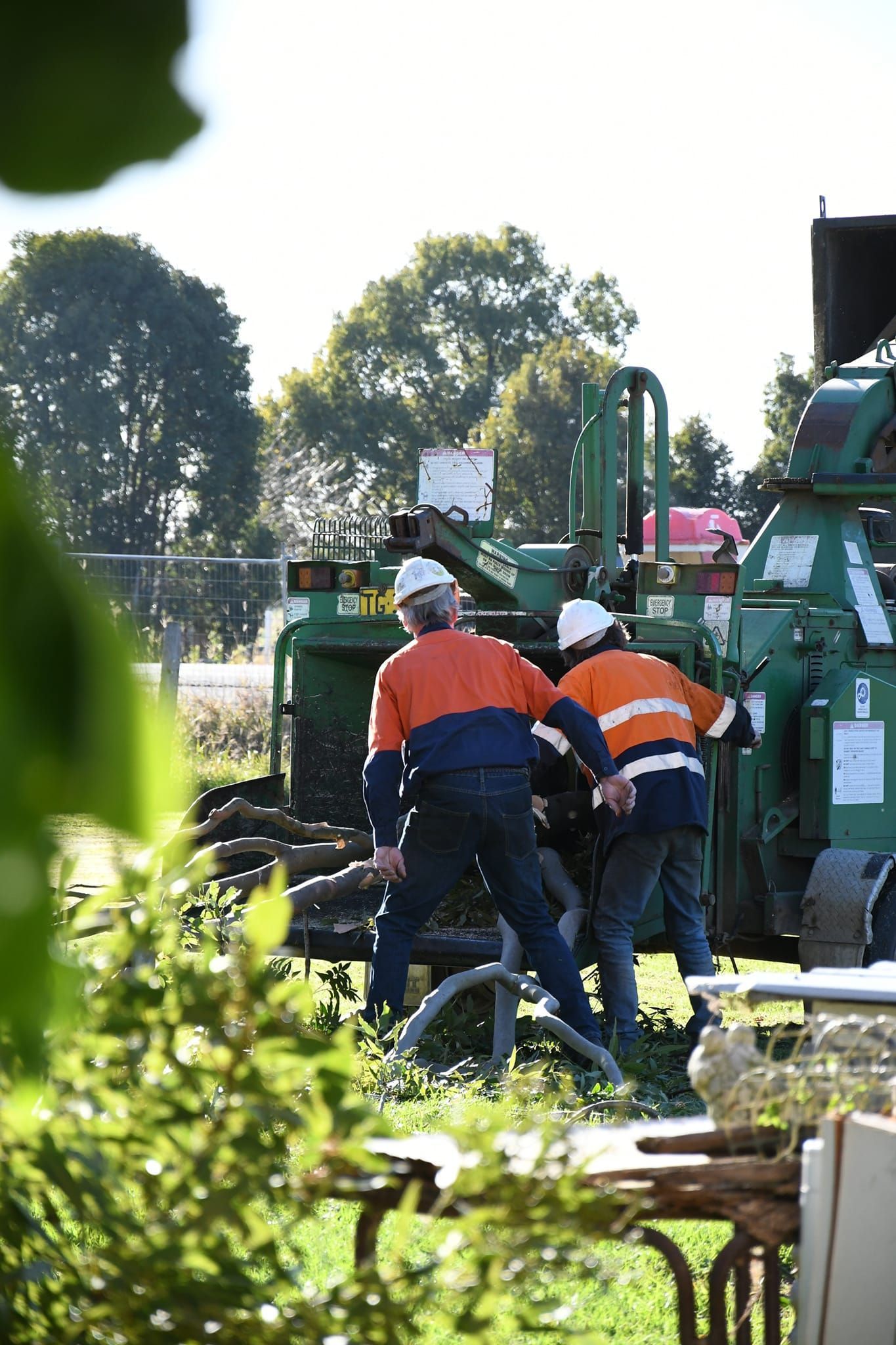Two workers in high-visibility orange and navy uniforms operate a large industrial wood chipper outdoors.