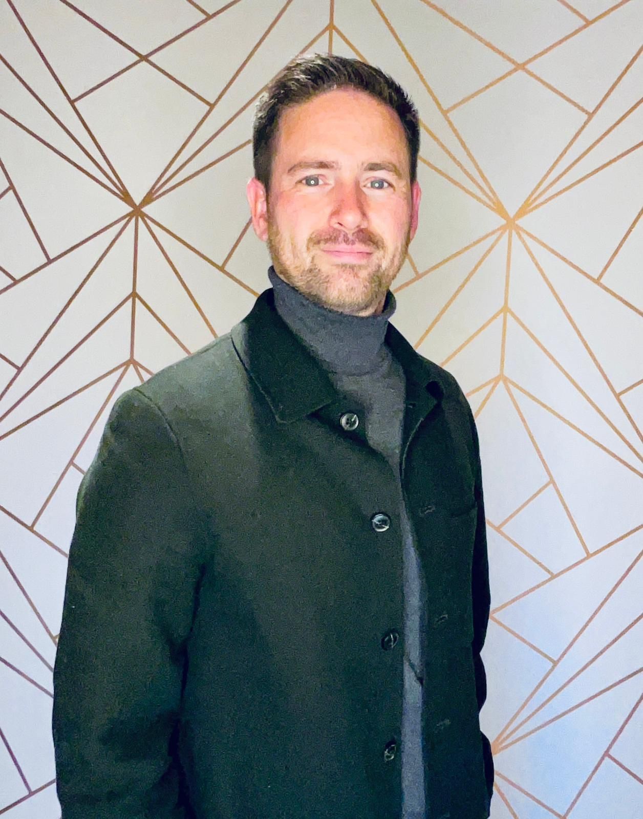 Een man in een zwarte jas en een grijze coltrui staat lachend voor een geometrisch muurpatroon.