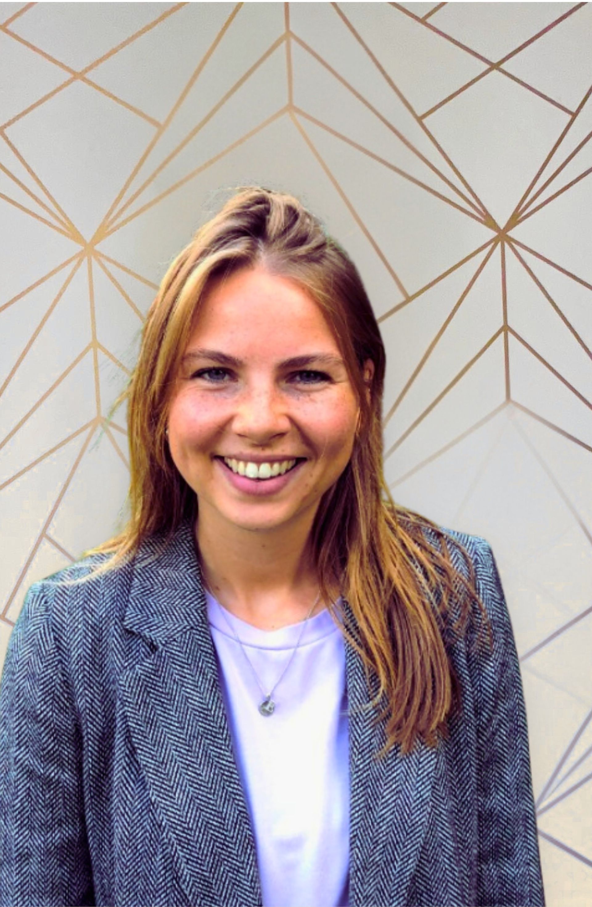 Een lachende vrouw, gekleed in een grijze blazer en een witte blouse, staat voor een muur met een geometrisch patroon.