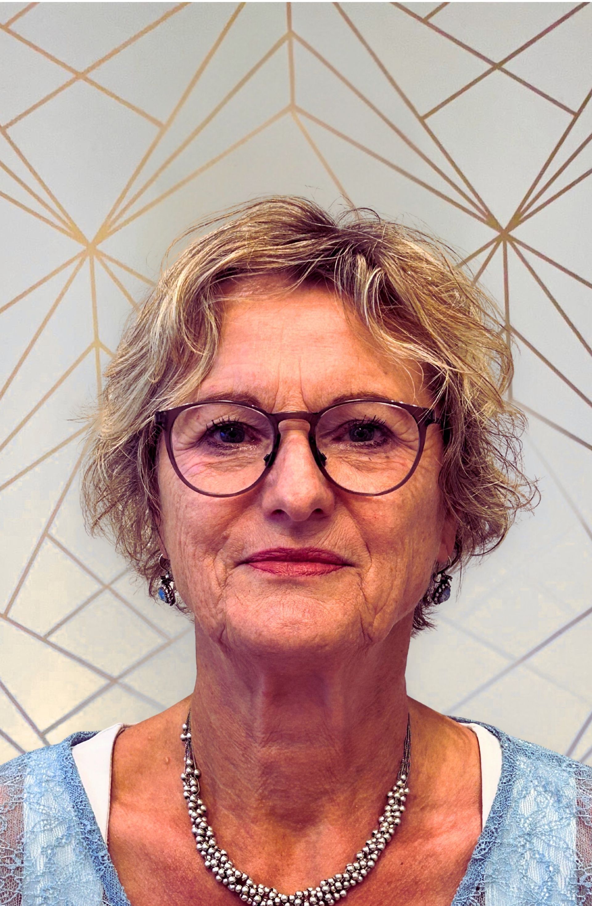 Vrouw met een bril en kort, krullend blond haar, draagt ​​een ketting en een lichtblauwe top.