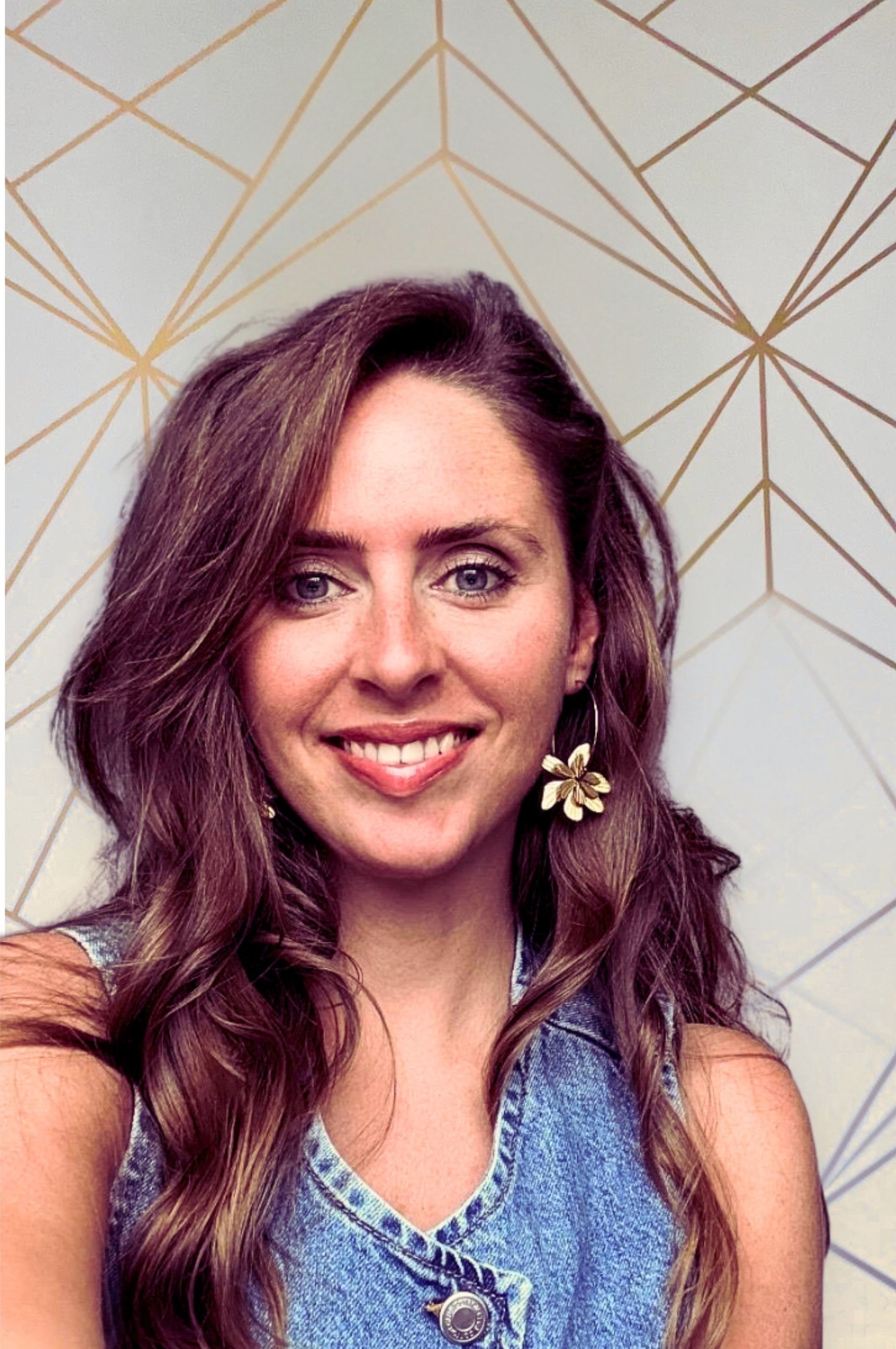 Vrouw met lang bruin haar, lachend, gekleed in een spijkerblouse en gouden bloemoorbellen. Geometrische achtergrond.