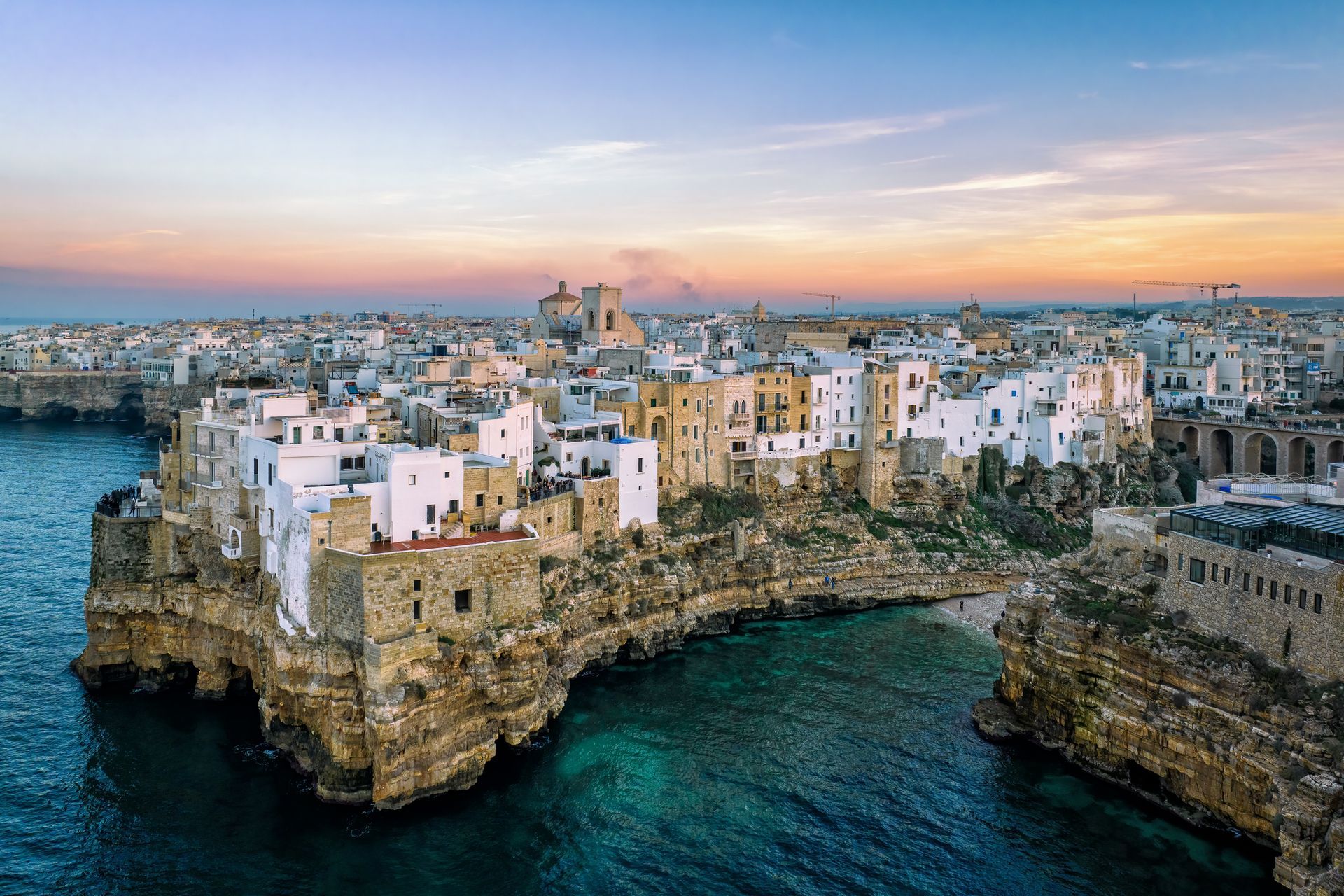 La cittadina costiera di Polignano a Mare, in Italia, con edifici bianchi arroccati sulle scogliere che si affacciano sull'acqua turchese al tramonto.