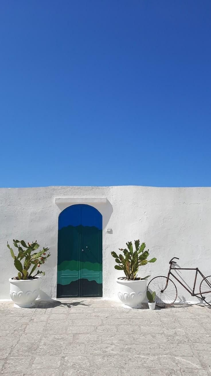 Muro bianco con porta blu e verde, piante di cactus in vaso, bicicletta; cielo azzurro brillante.