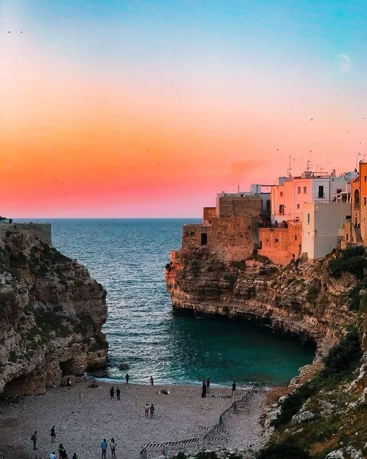 Città costiera al tramonto: edifici bianchi sulle scogliere si affacciano su una baia turchese e una spiaggia. Cielo arancione e rosa.