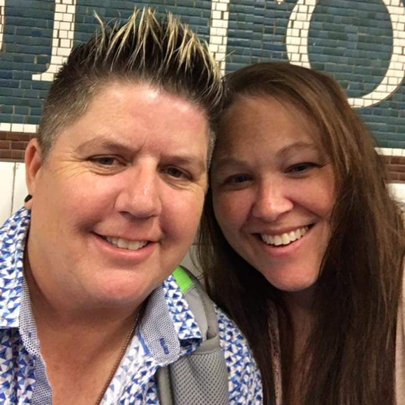 Two people smiling together in a close-up selfie in front of a blue tiled wall.