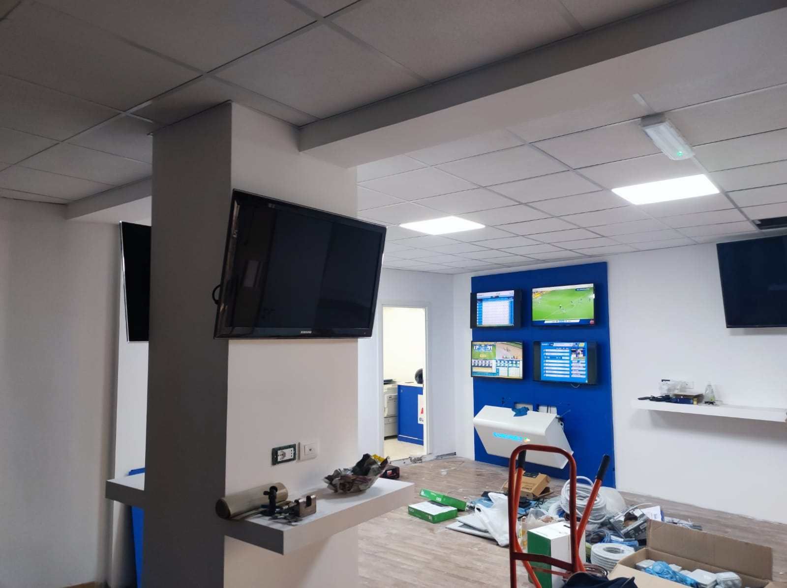 Interno di una stanza con più TV montate, una parete con accenti blu, detriti edili e una porta.