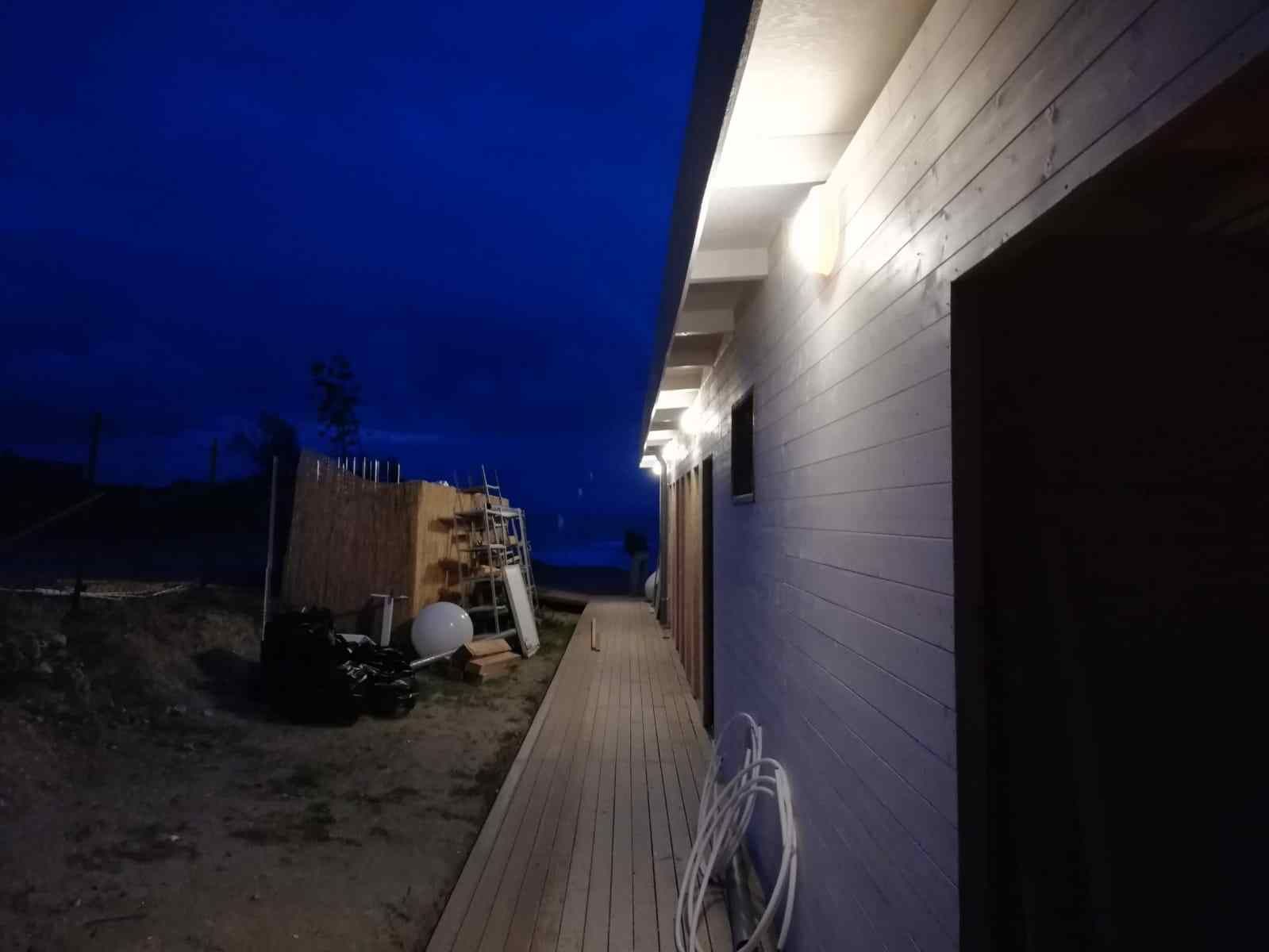 Vista notturna: una passerella di legno accanto a un edificio con luci accese. Cielo blu scuro.