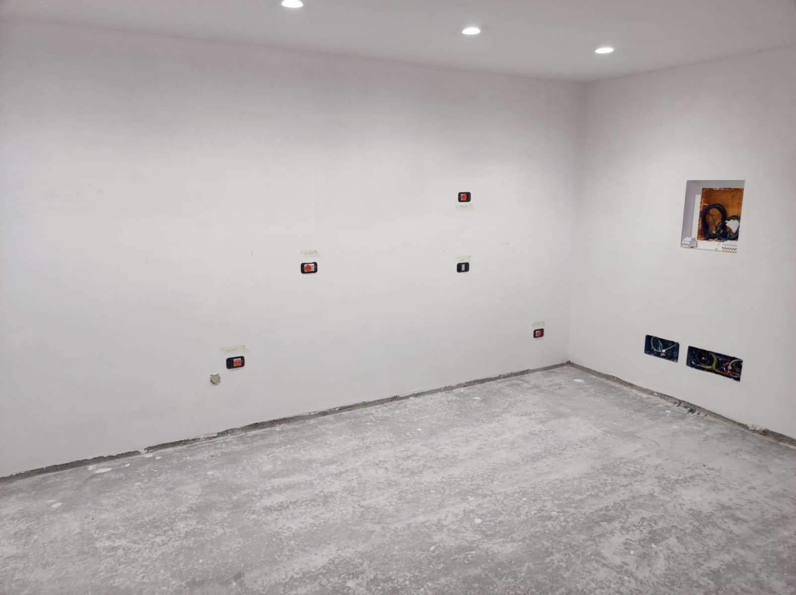 Stanza vuota con pavimento grigio, pareti bianche con prese elettriche e illuminazione da incasso.