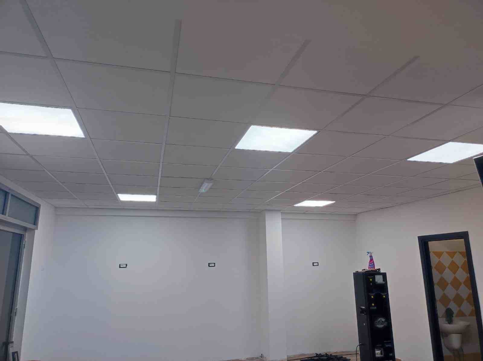 Una stanza con pareti bianche e un soffitto a griglia di luci fluorescenti. Una porta e una finestra si trovano sulla sinistra, mentre un armadio e una porta sono sulla destra.