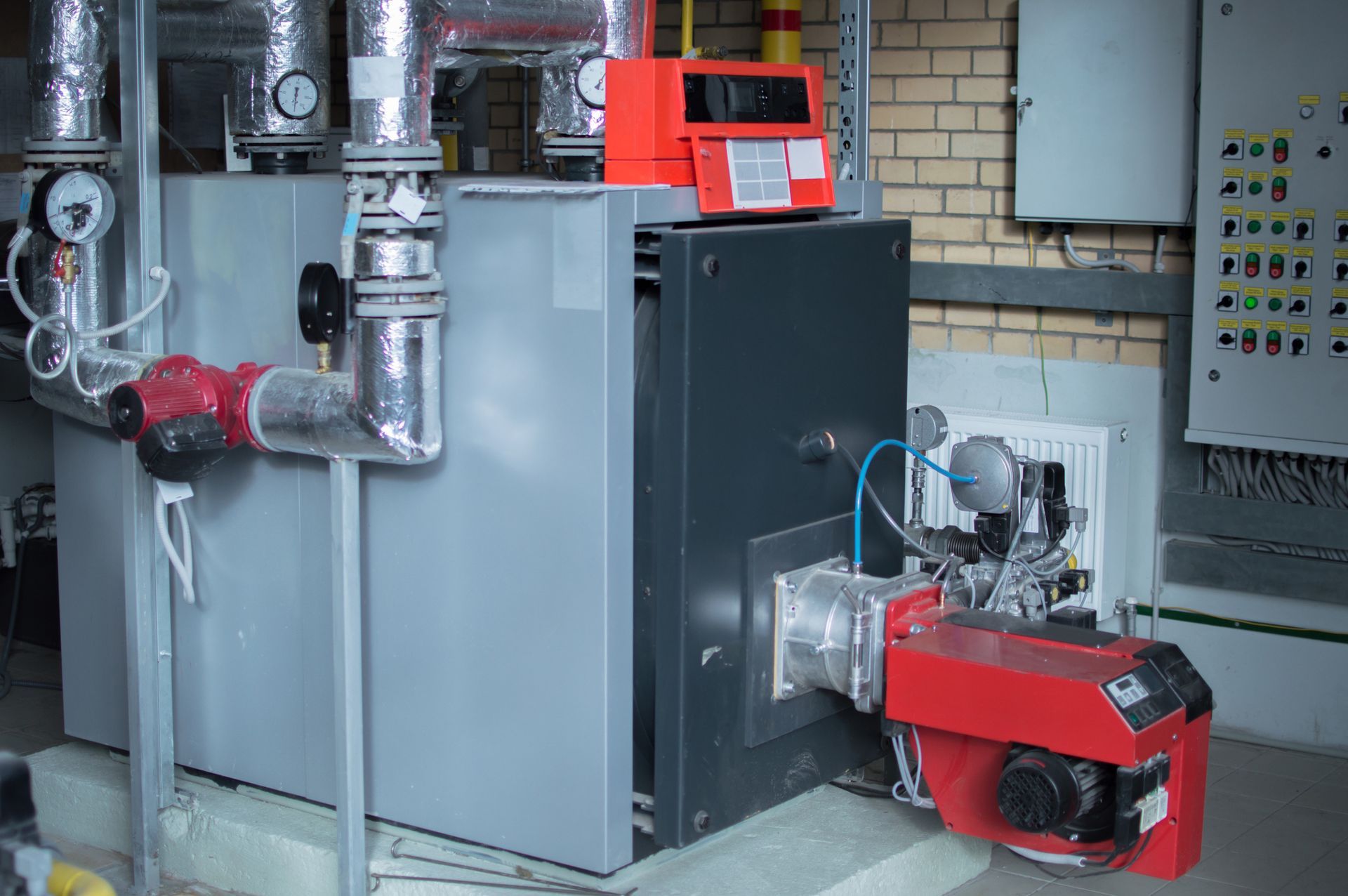 Caldaia industriale con bruciatore rosso, tubi, manometri e pannello di controllo in una sala macchine.