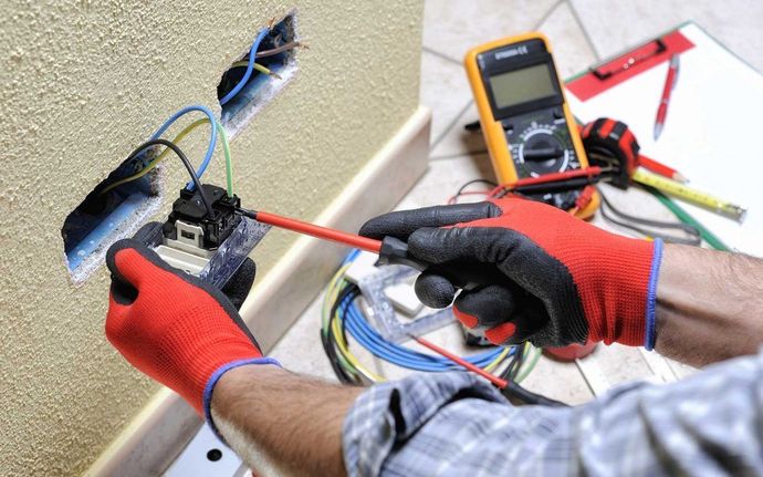 Elettricista con guanti rossi che collega i cavi di una presa a muro con un cacciavite.