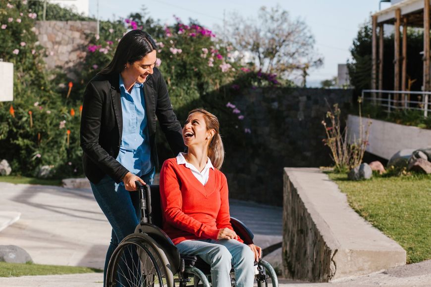 Woman in wheelchair being pushed by another woman, both smiling. Outdoors, sunny, near a building and greenery.