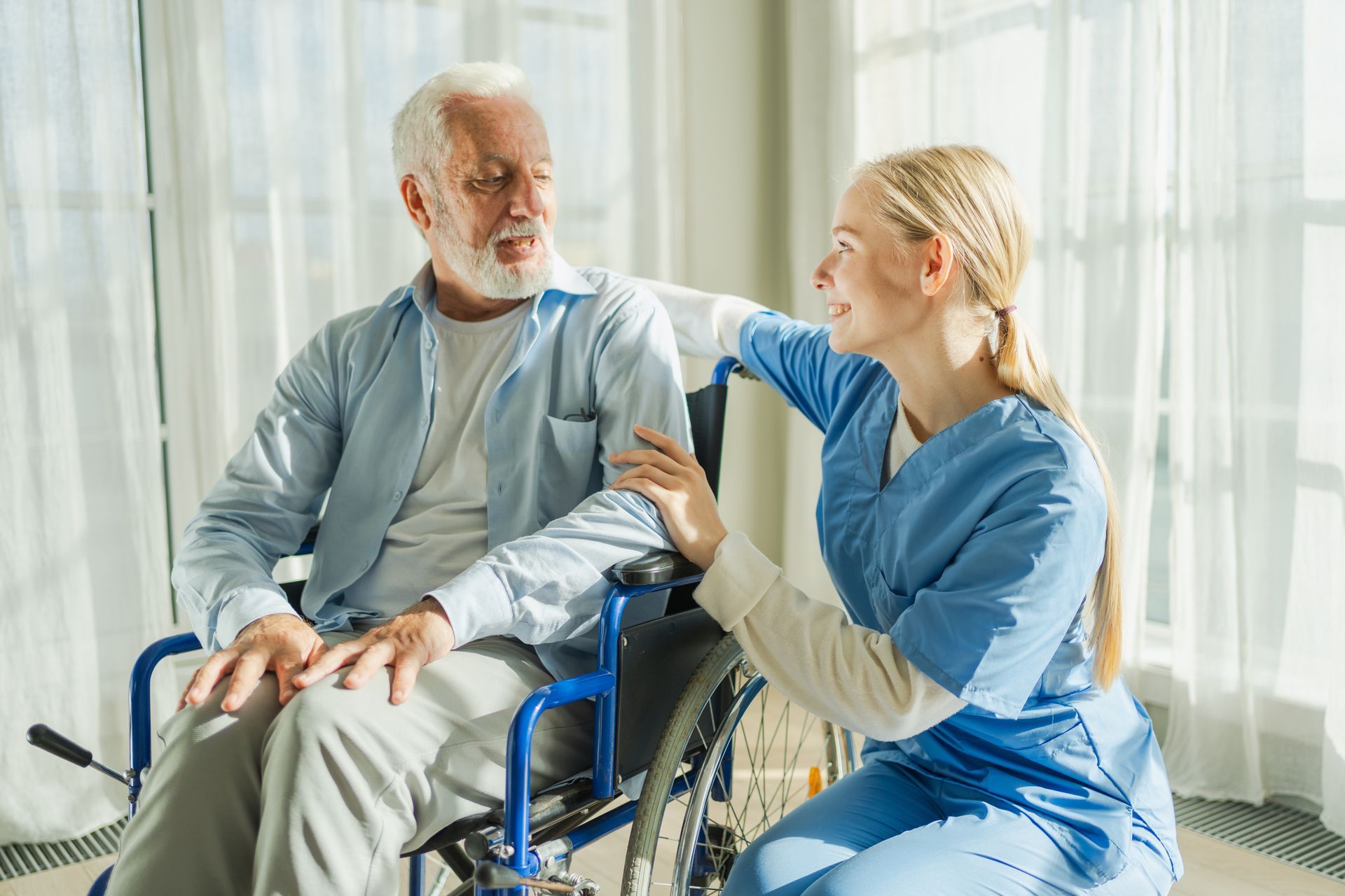 Elderly man in wheelchair talking with a smiling female healthcare worker in a bright room.