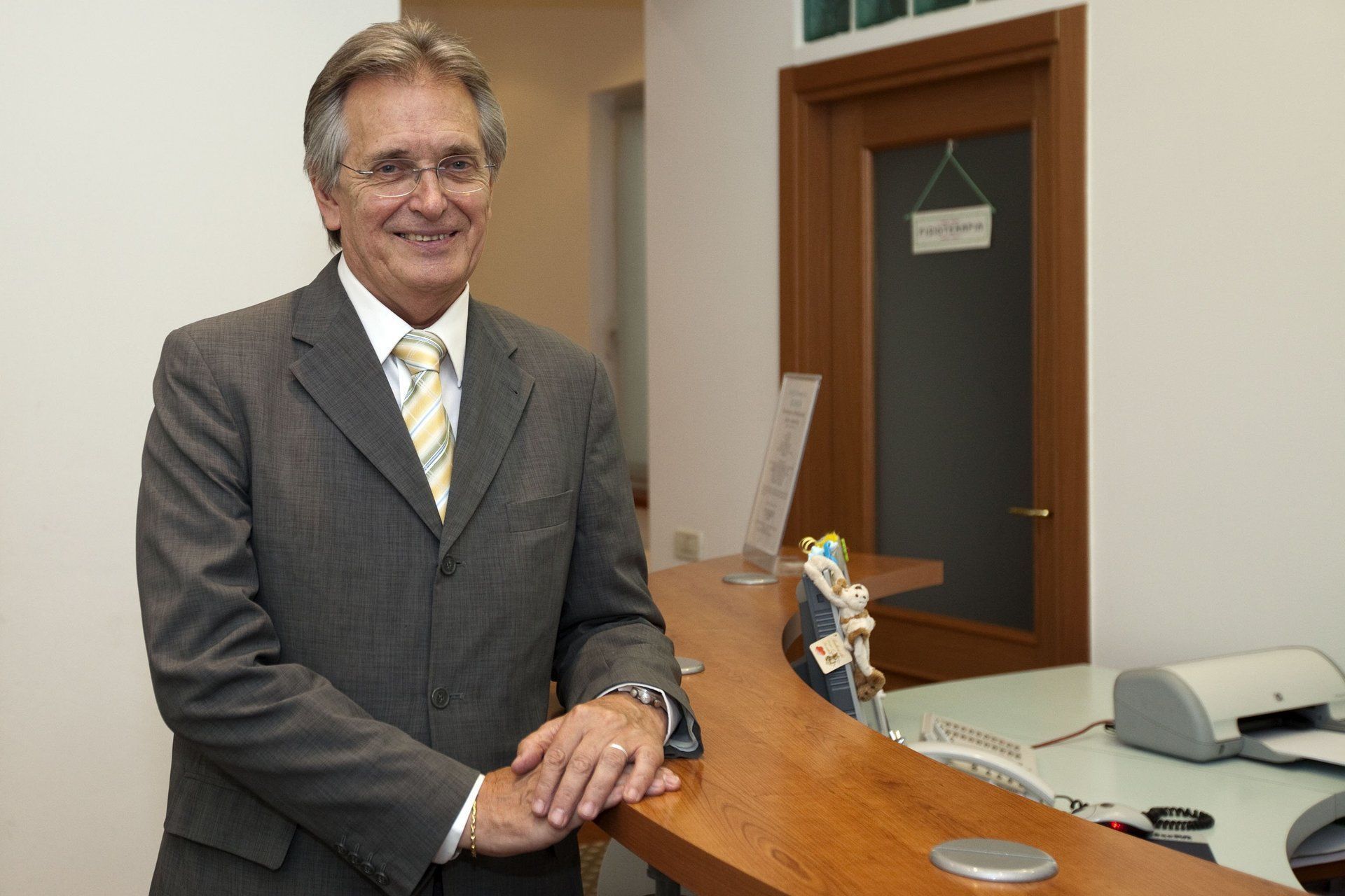Un uomo in giacca e cravatta sorride, in piedi dietro la reception di un ufficio.