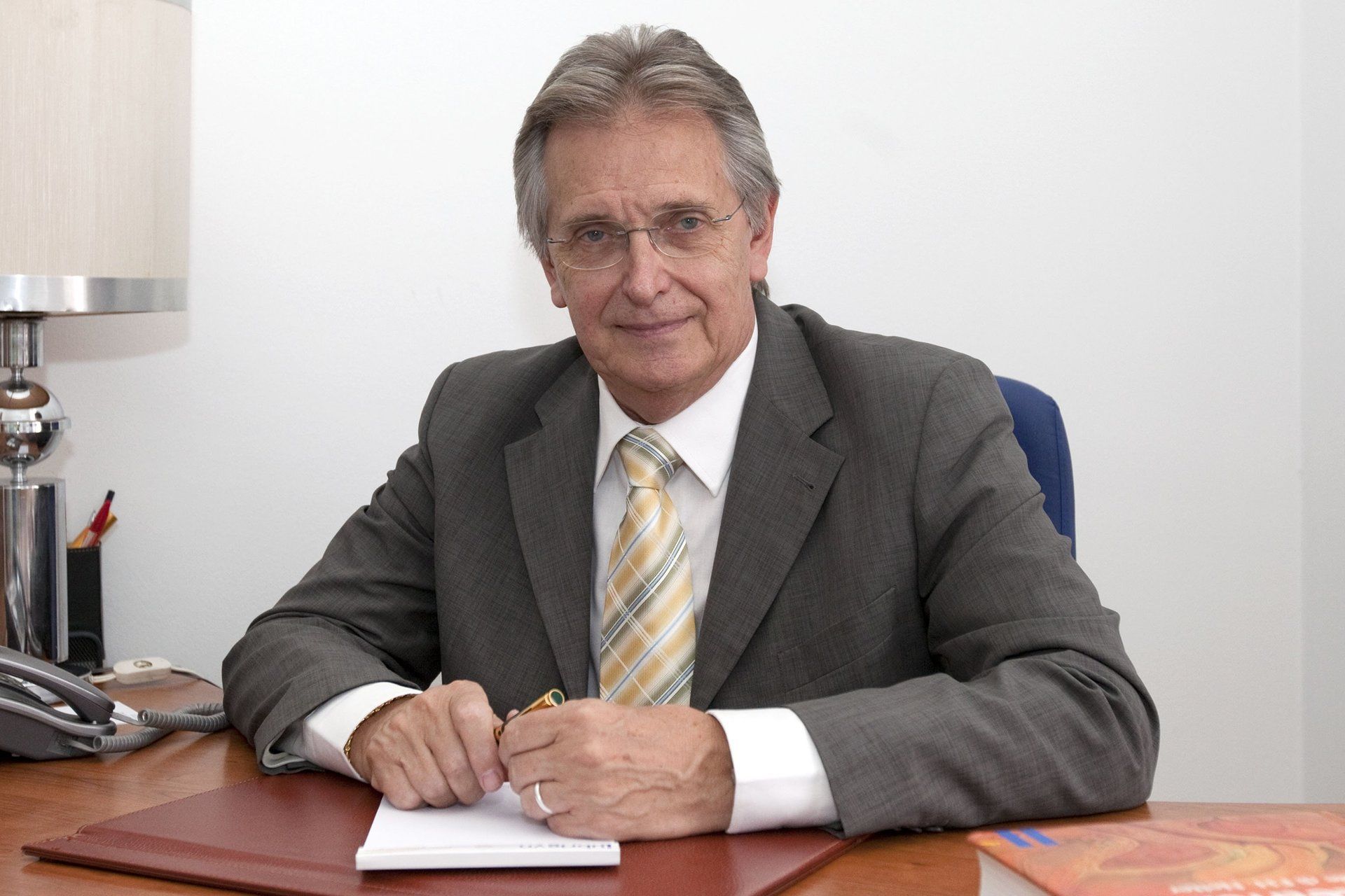 Uomo con occhiali, abito grigio e cravatta, seduto a una scrivania, che guarda la telecamera.