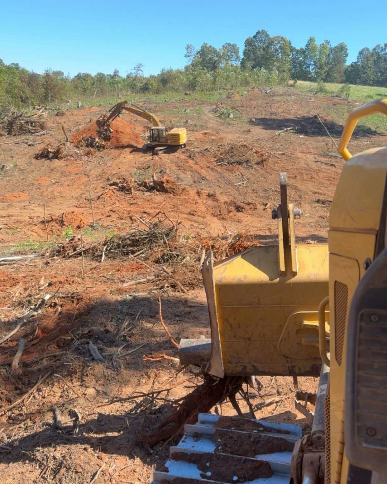 Yellow bulldozer in the foreground; excavator clearing land in background. Brown earth, blue sky, trees.