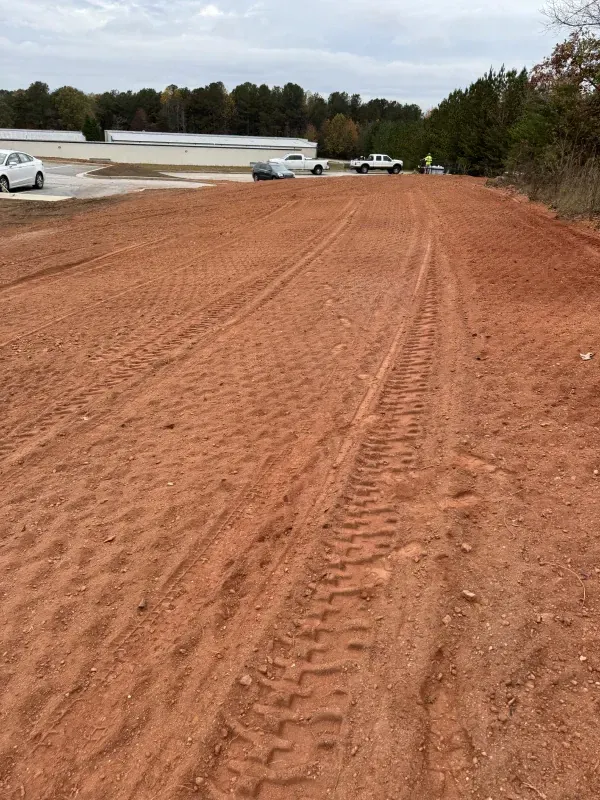 Red dirt lot with tire tracks, vehicles, and a building under a cloudy sky.