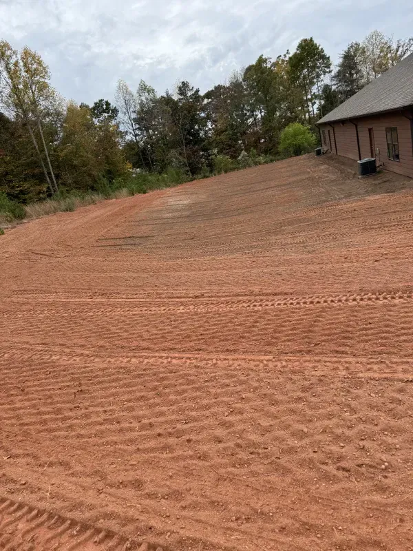 Red soil graded on a sloping lot, near a brick building and trees, under a cloudy sky.