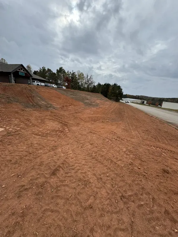 Red dirt hillside leads to water with trees and cloudy sky. A building is visible on the left.