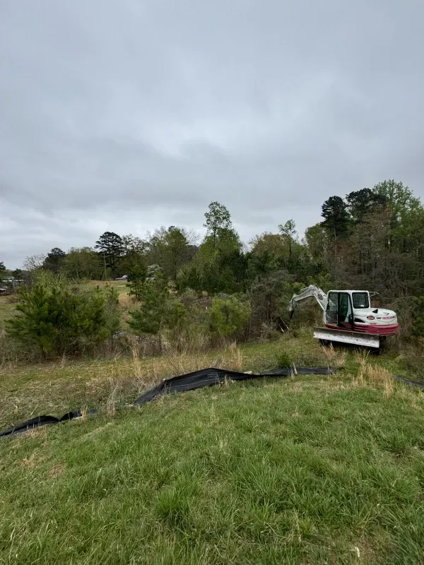 An excavator clears brush in a grassy field on a cloudy day.