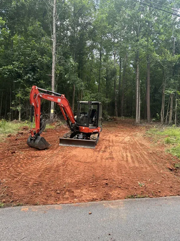 An orange Kubota excavator on a dirt plot cleared in a wooded area, next to a paved road.
