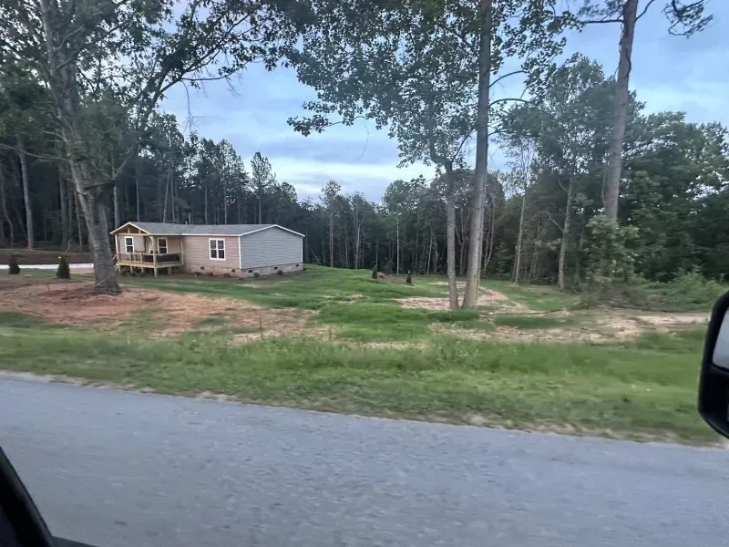 A small, light-colored house with a porch sits in a clearing, surrounded by trees and a grassy yard.