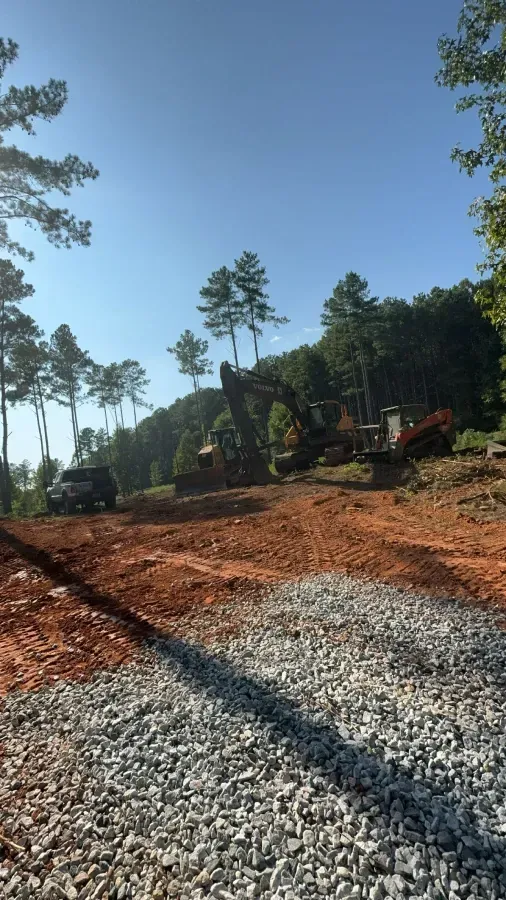 Construction site with excavator and truck on a dirt road, trees in the background, blue sky overhead.