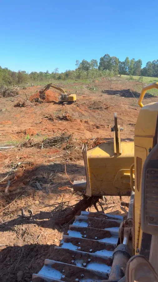 A yellow bulldozer clearing red dirt on a construction site; excavator in the distance.