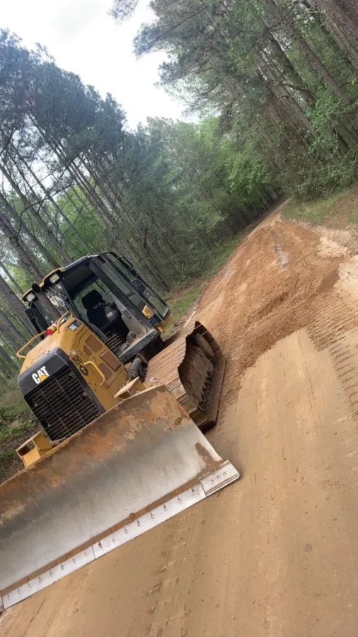 Bulldozer leveling dirt path in a wooded area. Yellow Caterpillar bulldozer is angled, pushing soil.