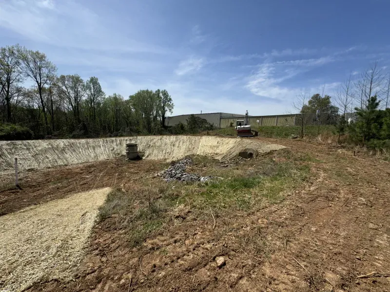 Construction site: excavated basin with gravel lining, dirt and grass, trees and building in the distance.
