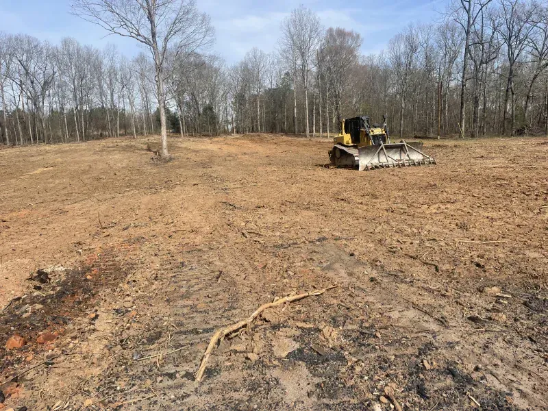 A bulldozer clears a brown field, trees in the background, under a blue sky.