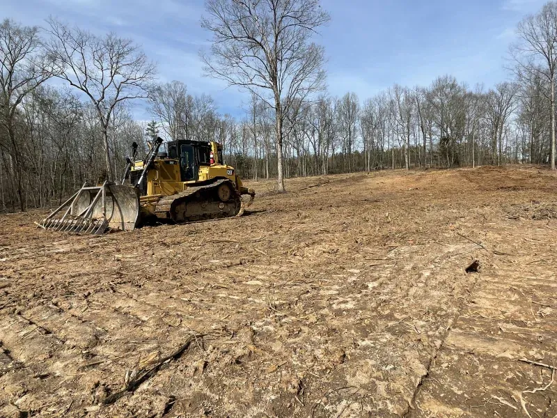 Yellow bulldozer clearing a brown dirt field on a sunny day. Trees in the background.
