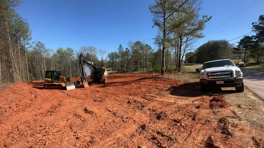 Construction site with heavy machinery on red soil, truck parked on side of road, trees in the background under blue sky.