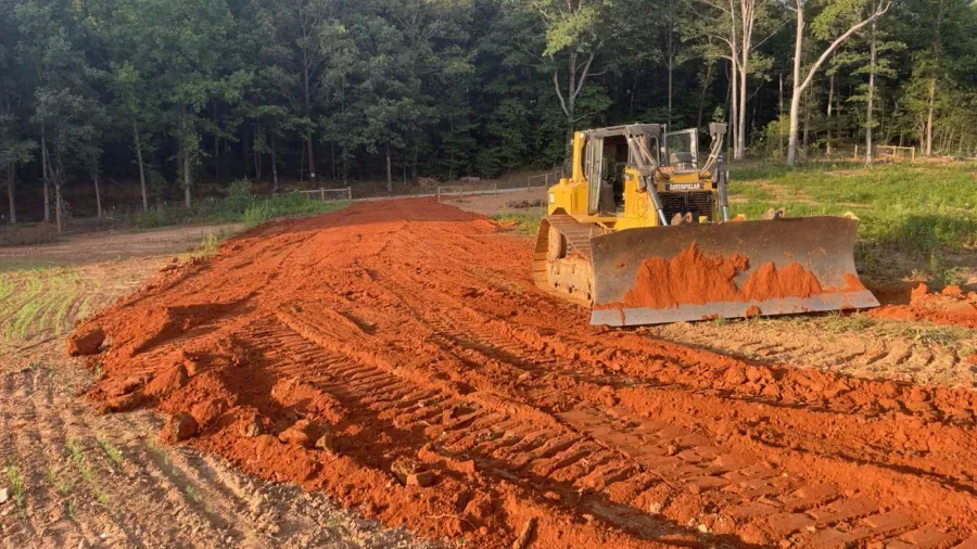 Yellow bulldozer pushing red soil on cleared land, with trees in the background.