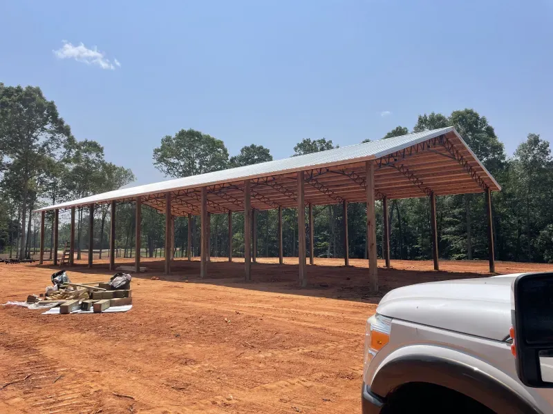 Open-air wooden structure with metal roof under construction; dirt ground; white truck in foreground; blue sky.