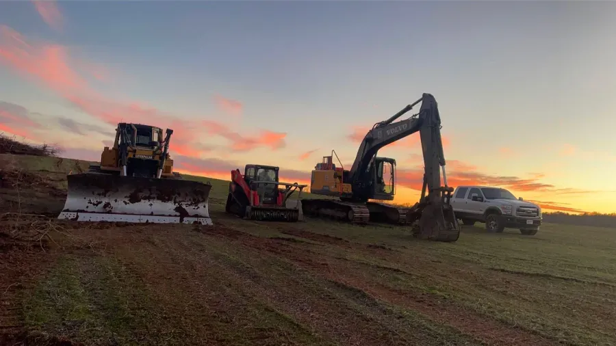 Construction equipment on a grassy field at sunset: bulldozer, excavator, skid steer, and truck.