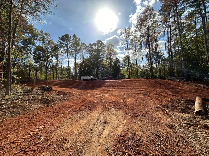 Cleared red dirt lot with trees in the background, under a bright sun.