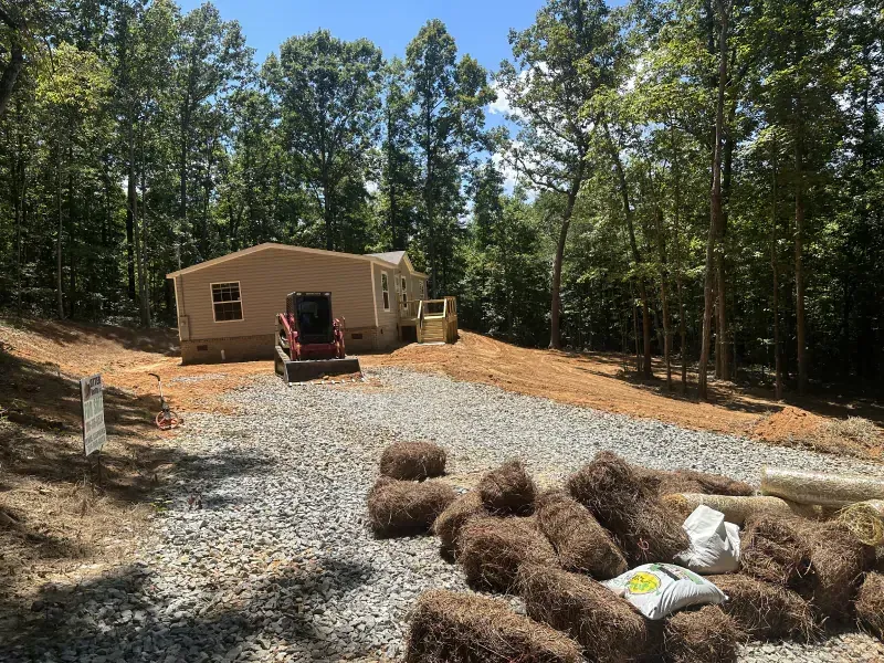 Mobile home on a gravel driveway with surrounding woods. Straw bales and construction equipment are present.