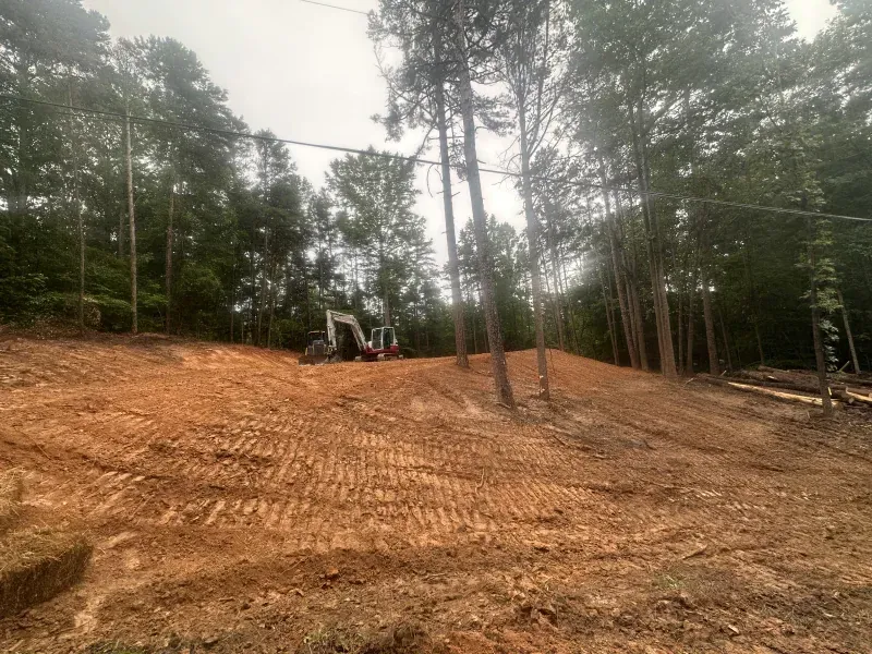 Earthmoving equipment on a leveled, reddish-brown dirt slope, trees in the background, overcast sky.