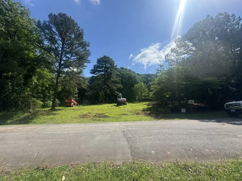 Cleared lot with construction equipment; road in foreground; trees in background; sunny.